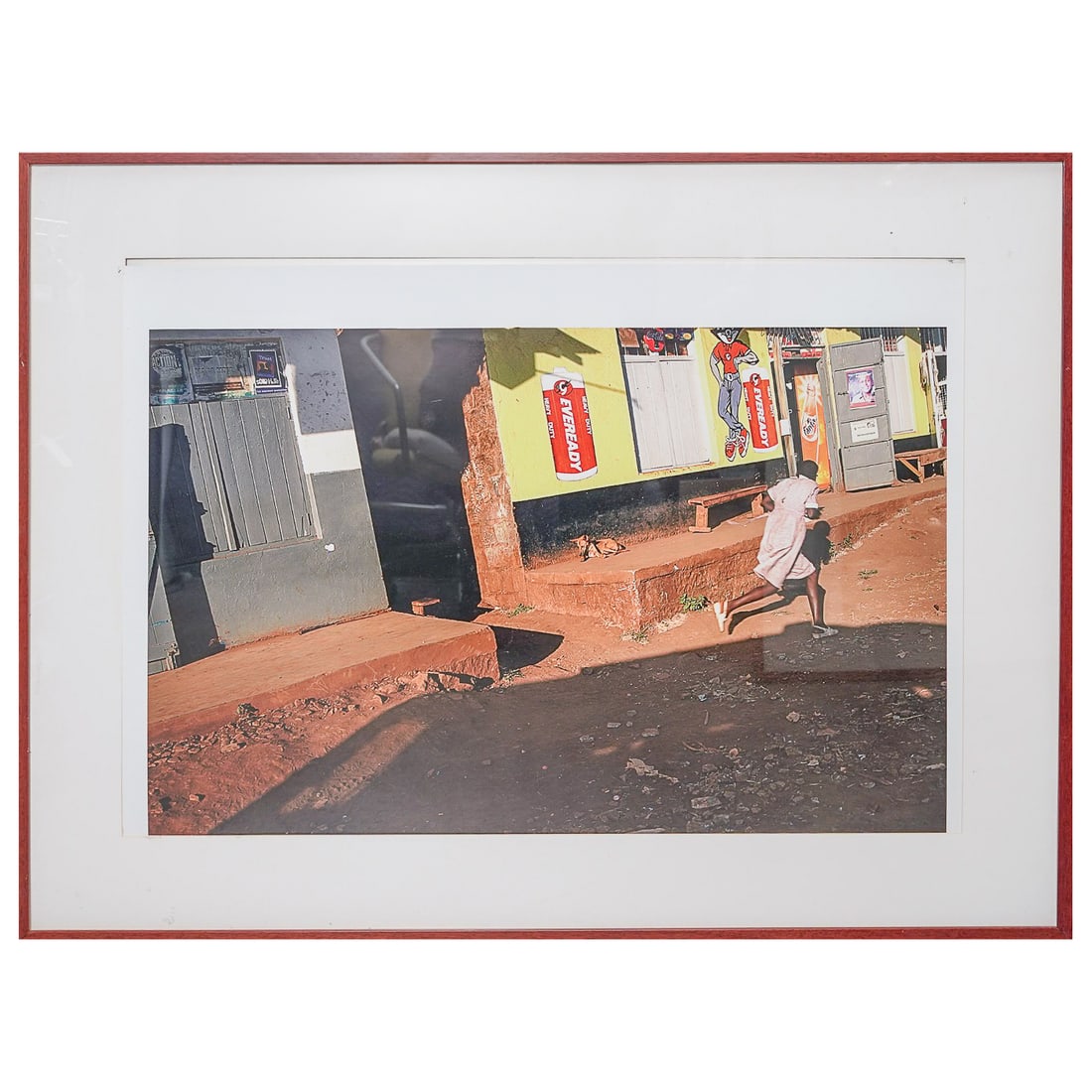 Attrib. Steve Mccurry Photograph Print: Attrib. Steve Mccurry Photograph Print20th century, KenyaPoster captures a moment in time, with a person in a pink dress running past the yellow and red storefront. The Eveready brand has been in the