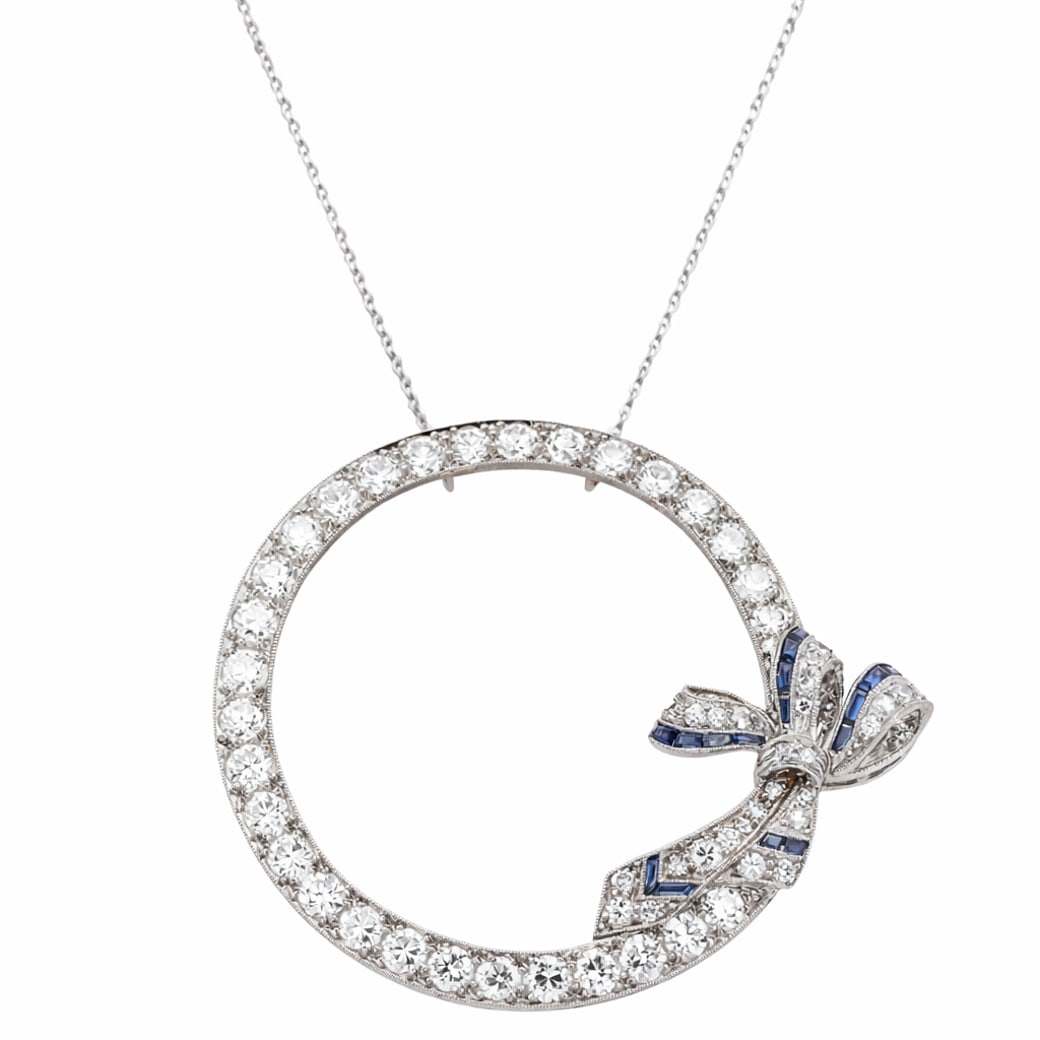 Art Deco Platinum Diamond & Sapphire Circle Bow Necklace: Art Deco Platinum Diamond & Sapphire Circle Bow NecklaceCirca 1930s-1940sCircular openwork pendant necklace executed in platinum, centering a diamond-set hoop accented by a stylized bow motif. The cir