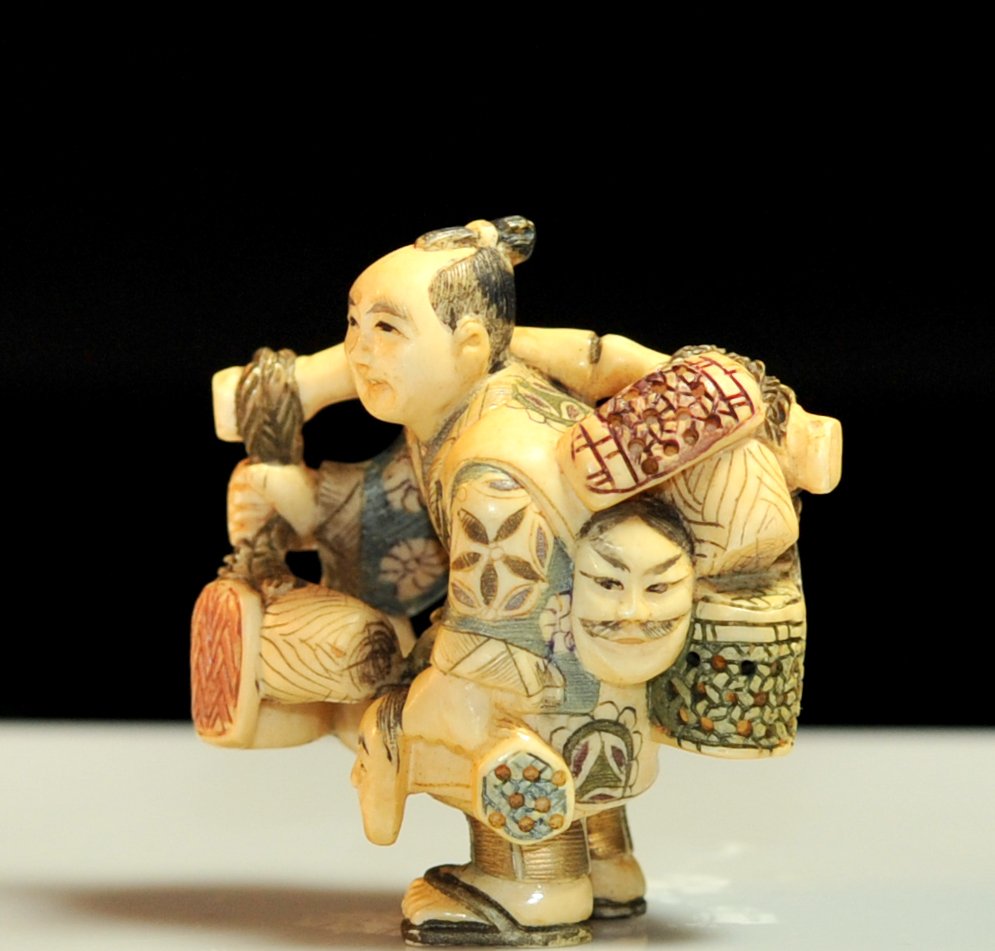 Chinese hand carved ivory "Entertainer" netsuke: DESCRIPTION: Chinese hand carved ivory "Entertainer" netsuke. Depiction of a robed man (One man show) who carries multiple masks and baskets draped over a large bamboo stick. Full polychrome designs t