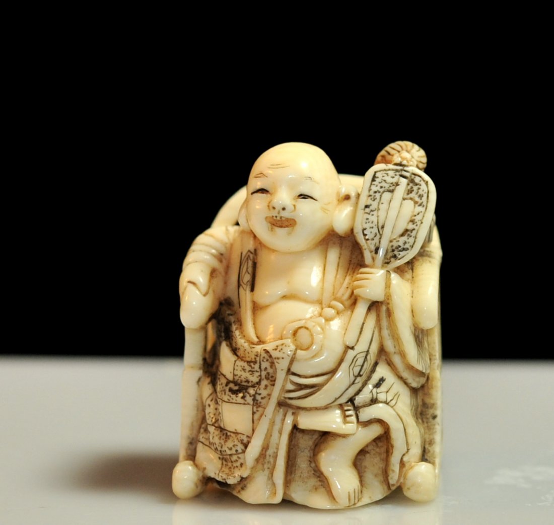 Vintage Chinese "Sitting Buddha" carved netsuke: DESCRIPTION: Vintage Chinese "Sitting Buddha" carved netsuke. Features the Buddha draped in a long flowing robe. He holds a fan in one hand with his other hand over the arm of the chair in which he si