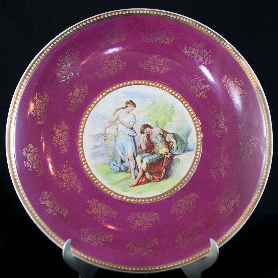 Red English Porcelain Charger: DESCRIPTION: A red charger with gold floral designs. In the middle there is a scene of a man sitting down and a standing woman. On the bottom of the charger is a hallmark from Empire Works. CIRCA:Mid