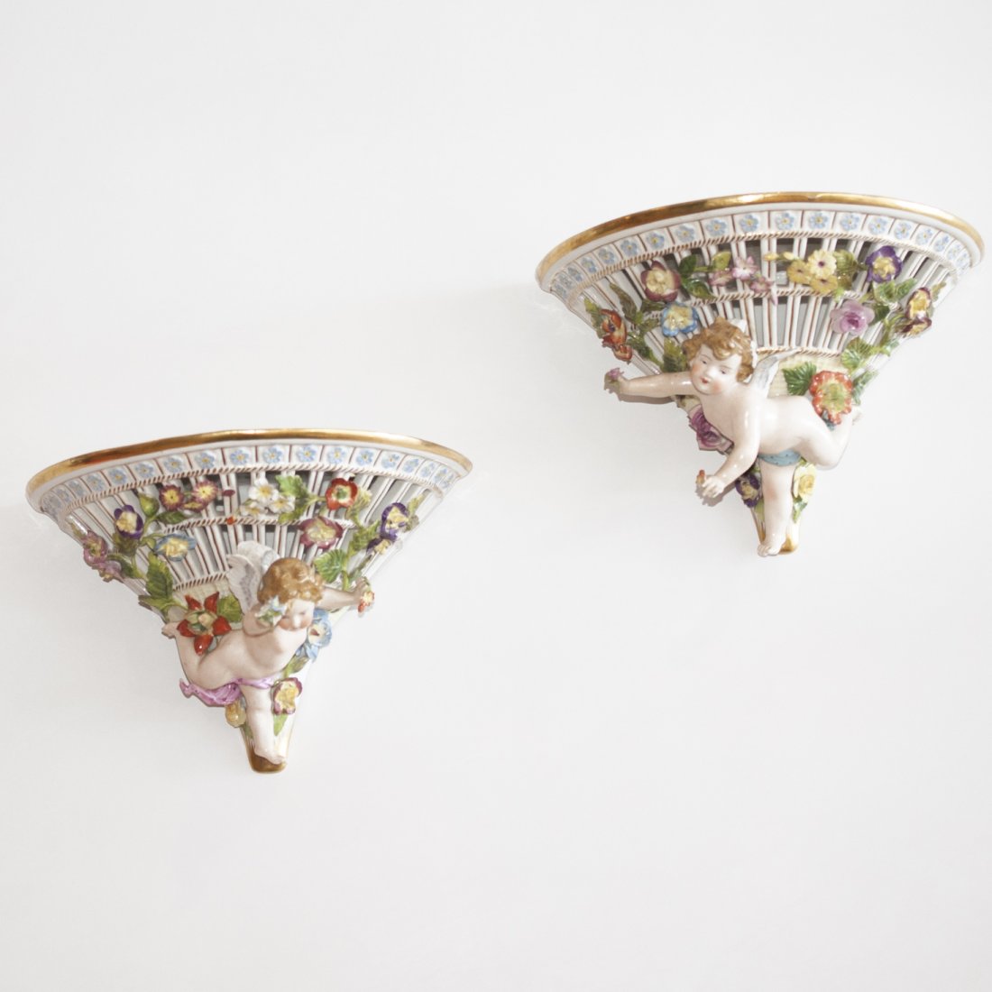Pair of Meissen Porcelain Sconces: DESCRIPTION:Pair of handpainted Meissen porcelain sconces. Features handpainted reliefs on affable cherubs gliding in front of a bed of flowers. Finished with a flat top with gilded trim and polish. M