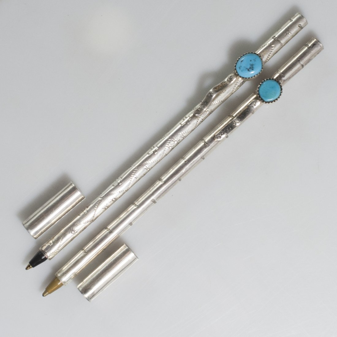 Sterling & Turquoise Pens: DESCRIPTION:Pair of sterling ballpoint pens. One features engravings of crosses, suns, and spirling design, the other features engraved "Sterling" marks along the exterior. Both are fitted with turquo