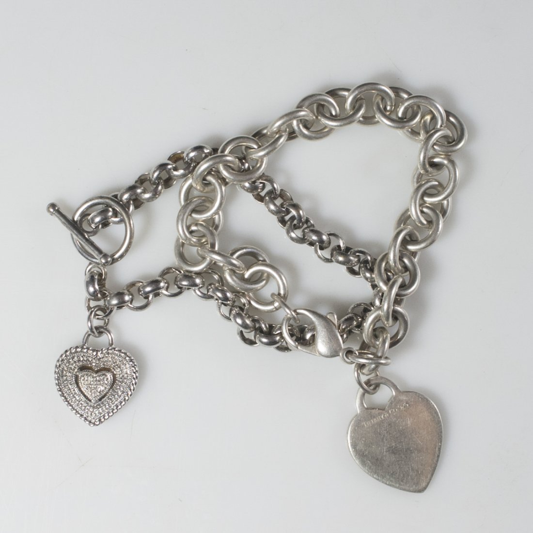 Tiffany & Co. & Sterling Heart Bracelets: DESCRIPTION:Pair of sterling bracelets. One is Tiffany & Co. and features a chain links that convene at a lobster claw clasp and finished with a heart shaped pendant marked:"Tiffany & Co. 925". The ot