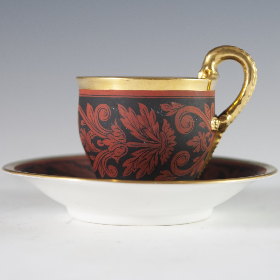 Antique KPM Porcelain Demitasse & Saucer: DESCRIPTION: An antique hand painted KPM porcelain demitasse and saucer. Decorated with winding rococo floral motif in black and red hues. Finished with gilded accents along the saucer and gilded inte
