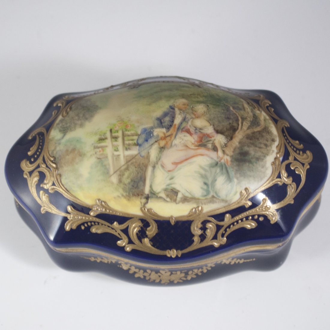 Antique Sevres Porcelain Trinket Box: DESCRIPTION: Antique Sevres porcelain trinket box. Features handpainted renderings of a Georgian era garden courtyard scene with a man leaning over a woman, backed by a garden and trees with bountiful