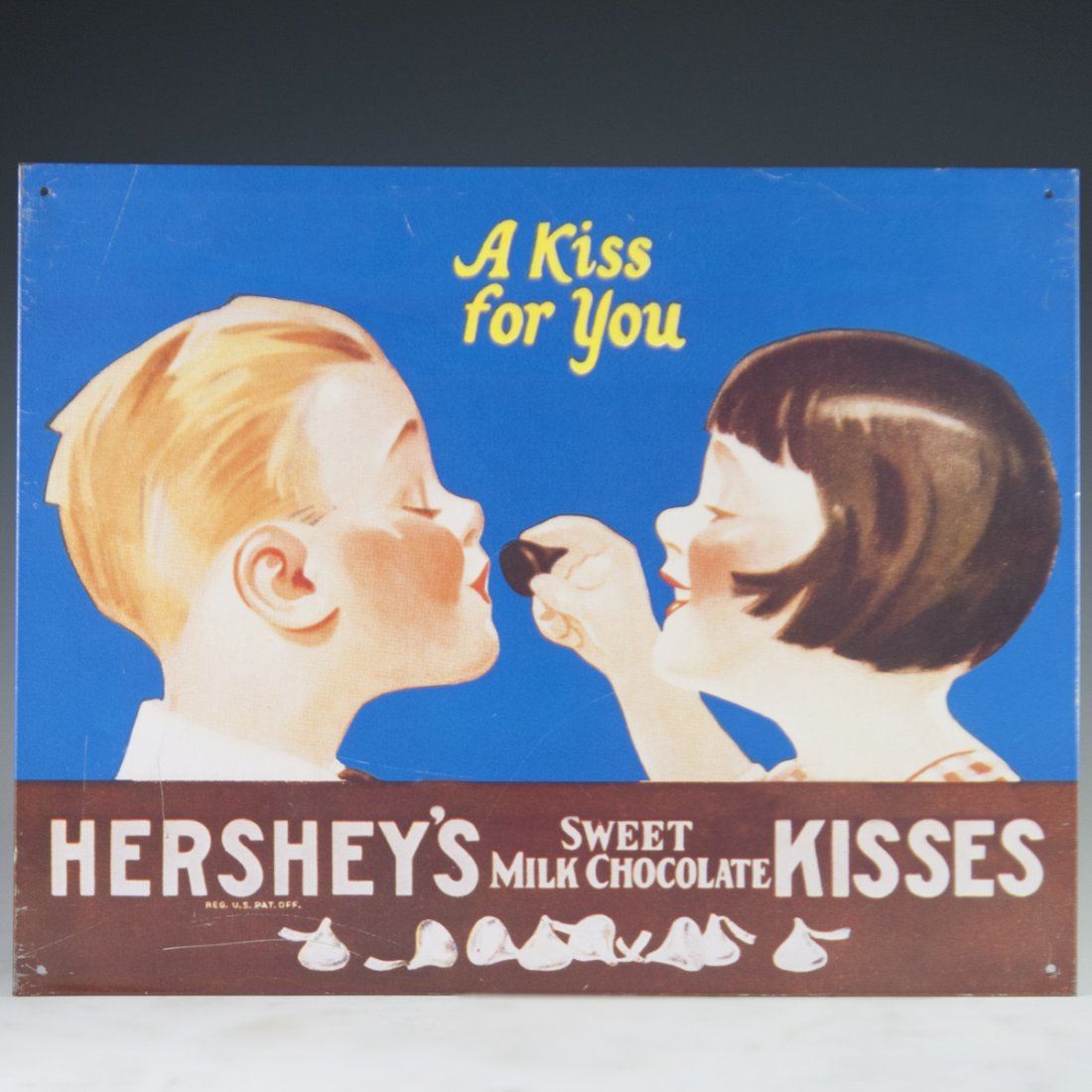 Hershey's Chocolate Aluminum Sign: DESCRIPTION: A Hershey's chocolate decorative aluminum advertisement sign. Features a rendering of a young girl feeding a boy a Hershey's Kiss. CIRCA: 20th Ct. ORIGIN: USA DIMENSIONS:H:12.5" L:16" CON