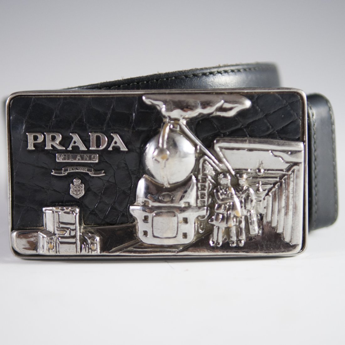 Vintage Prada Belt Buckle: DESCRIPTION: A vintage Prada belt buckle. Features a scene of a train arriving at a platform with figures boarding, backed by leather base. Marked:"Prada Milano DAL 1913". CIRCA: 20th Ct. ORIGIN: Ital