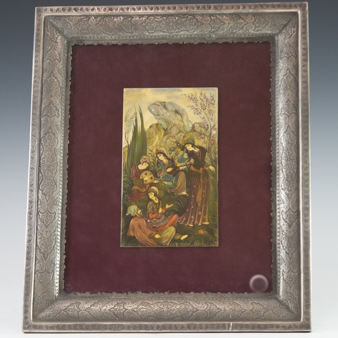 Persian Silver Framed Mughal Painting: DESCRIPTION: A Persian Mughal painting on bone featuring a rendering of regal figures, seated in a garden as they dance and listen to music. Finished with a scenic mountainous landscape backdrop along