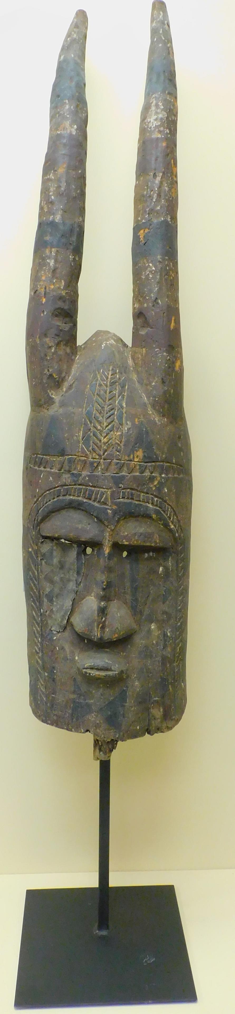 Large early 20th century carved African mask depicting: a horned figure, Bobo people. Remnants of old paint and bug damage. Mounted on modern metal base. Overall height with base 83 1/2". Mask is 62" high. Approx. 14" diameter.