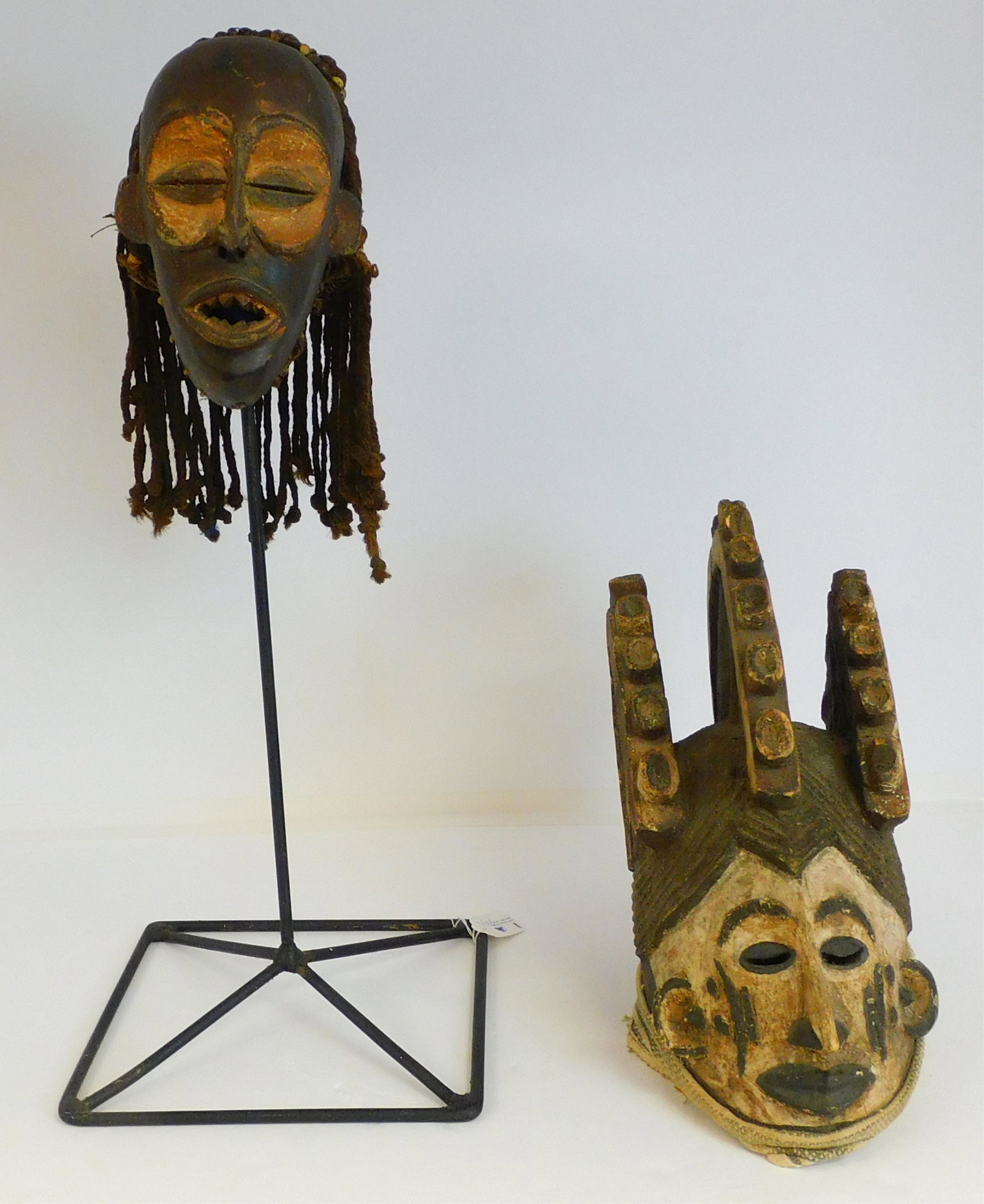 Two 20th century African masks. To include: a carved: mask with rope dreadlocks, Dan people, 8 1/2" high, approx. 6" wide, on a custom metal stand. Along with a carved and painted mask of a male figure, old worn polychrome paint, 14" high, 15" deep, 7" w