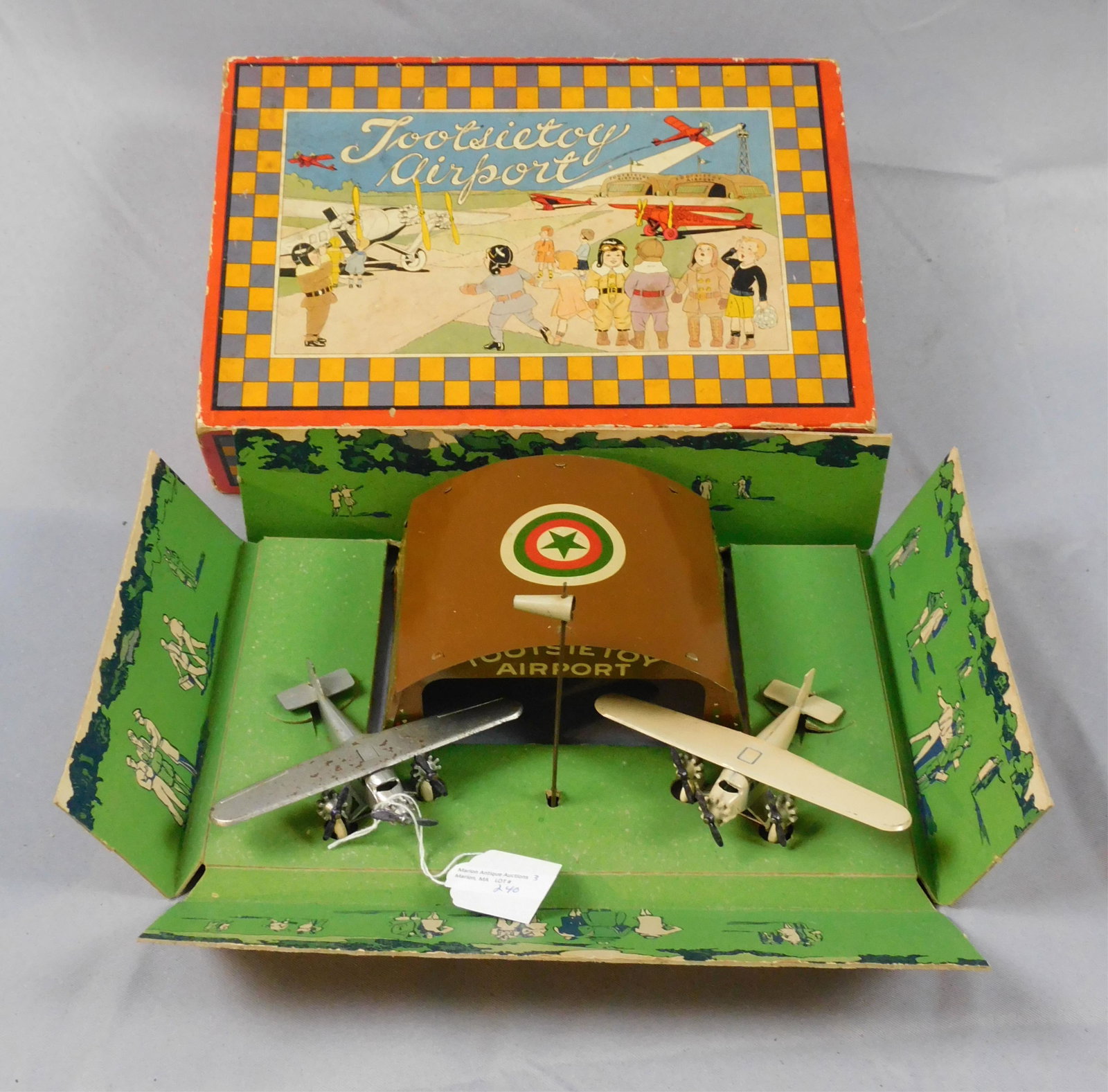 Tootsietoy Airport, original box with colored: lithograph cover, ca. 1930s. Contains two litho tin airport buildings with two planes. One plane shows paint loss to the top and wings. Box is 12" x 8" x 3".