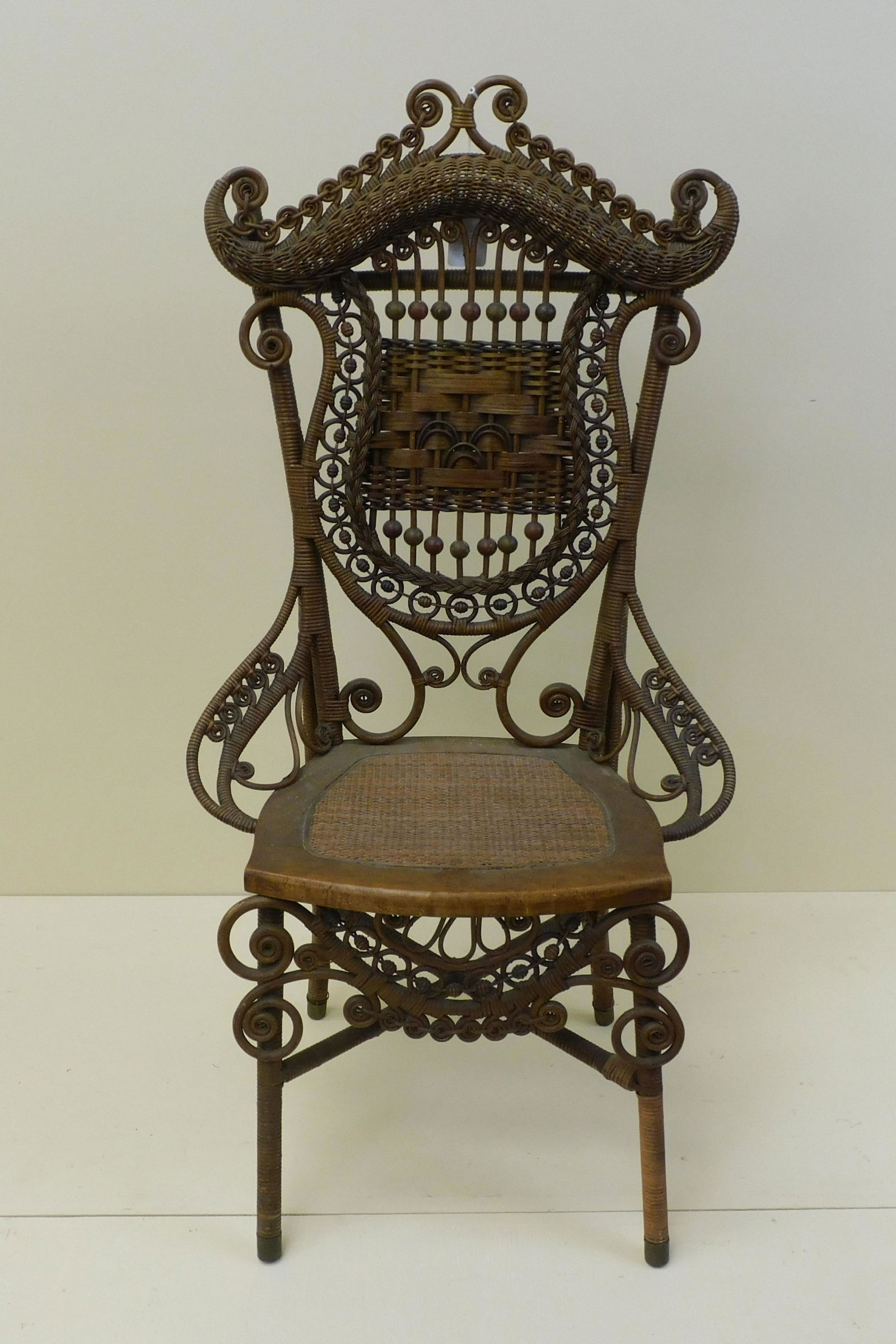 Elaborate wicker chair, probably by Wakefield: Rattan Company, 1890. Made in Wakefield, Massachusetts. The chair has a curlicued and rolled crest with a harp/shield shaped back. Maple seat frame and caned seat with brass feet. There are remnants o