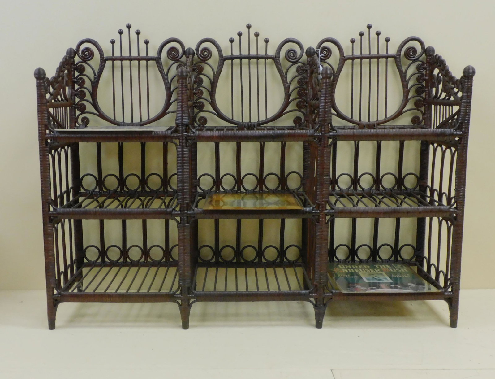 Victorian wicker music stand, ca. 1890, with: three divided sections, each having three shelves. Lyre design in back with ornate curlicue gallery sides. Dark natural finish is not original. 36" high, 48" wide, 15" deep. Good condition overall. Co