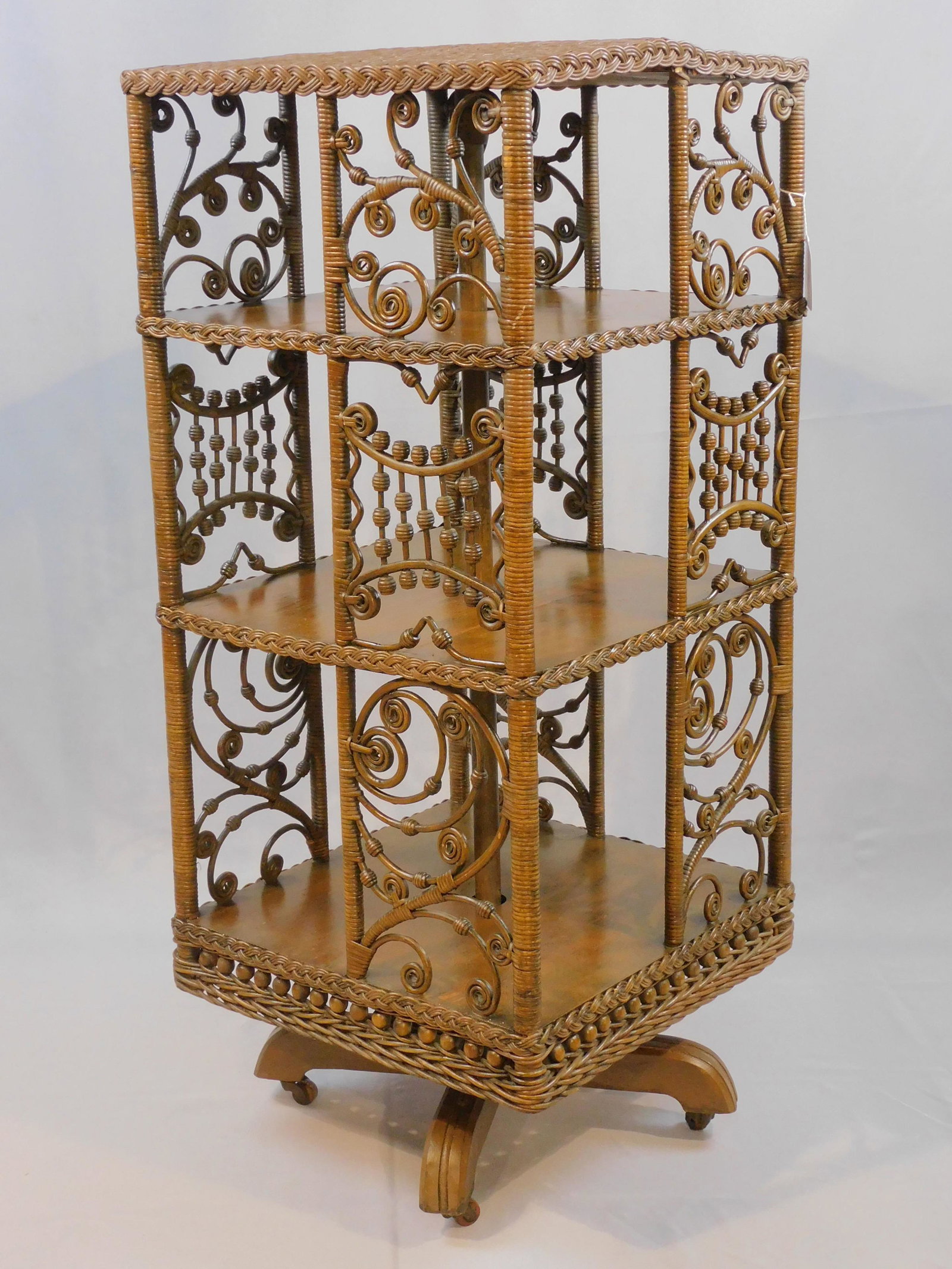 Victorian wicker revolving bookcase, ca. 1895.: Rattan, oak, and iron. Original natural finish. 3 tiers with decorative curlicue side panels. Wooden feet with casters. Very good condition. Top is slightly warped. 42" high, 18" wide, 18" deep. Prove