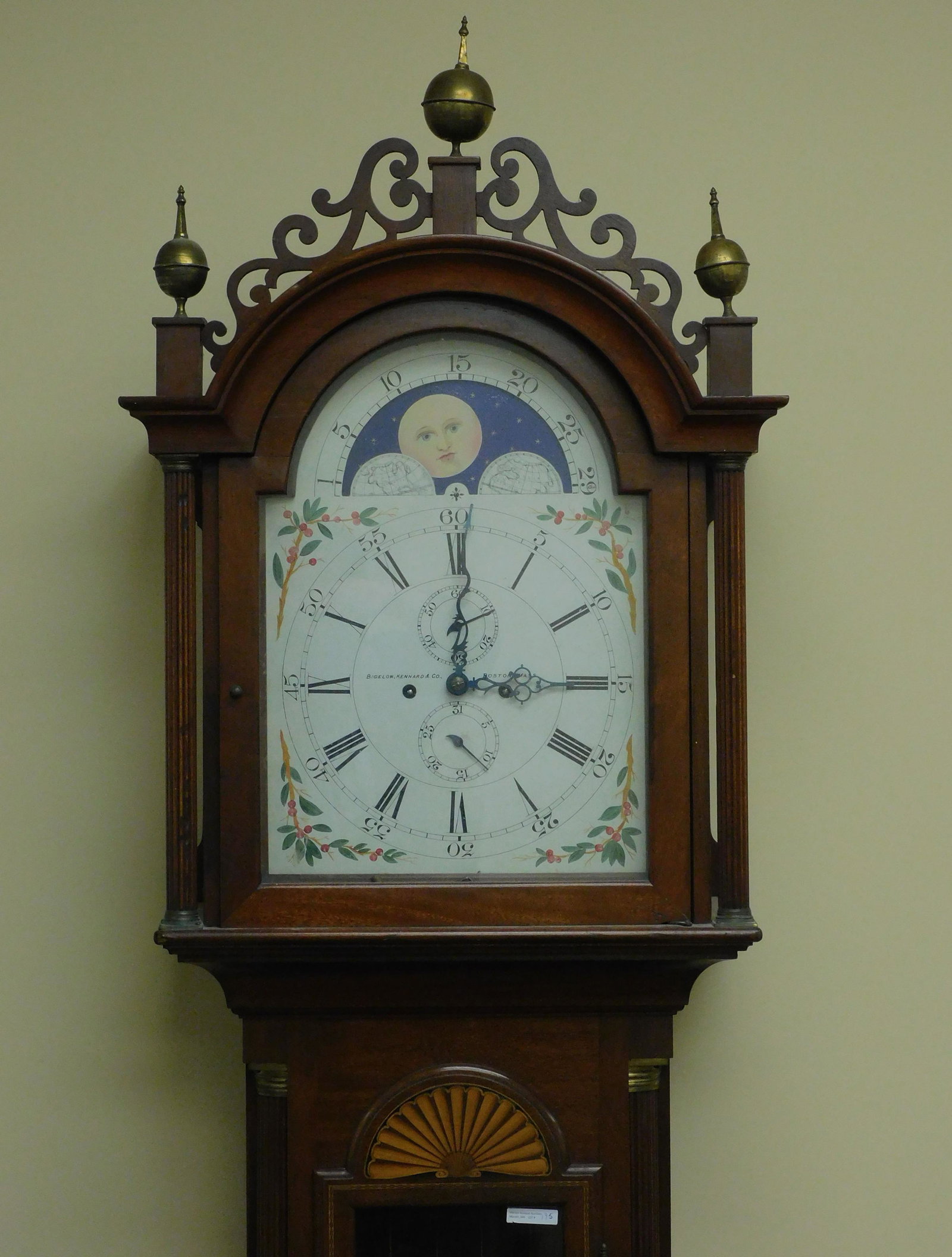Mahogany grandfather clock, early 20th century.: Cut out fretwork gallery and brass finials. Painted metal moon phase dial with calendar in seconds hand, signed Bigelow and Kennard Company, Boston, Mass. Central door has glass panel and inlaid fan d