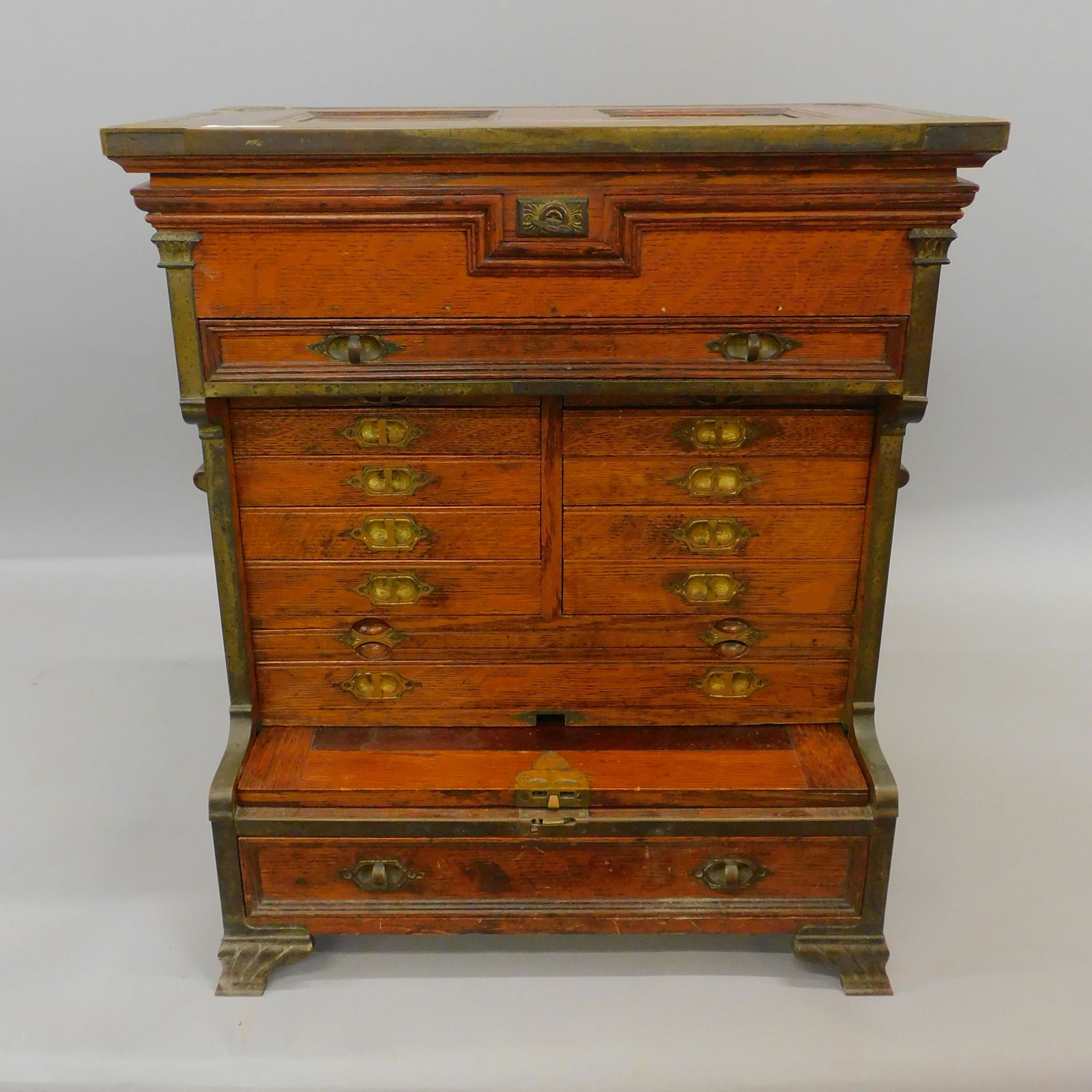 An exceptional quartered oak machinist chest with: ornate brass trim. Lift top with two trays over a thin single drawer over eight drawers over two long drawers, pullout writing surface over one large drawer. Ornate large bracket feet. Internal brass