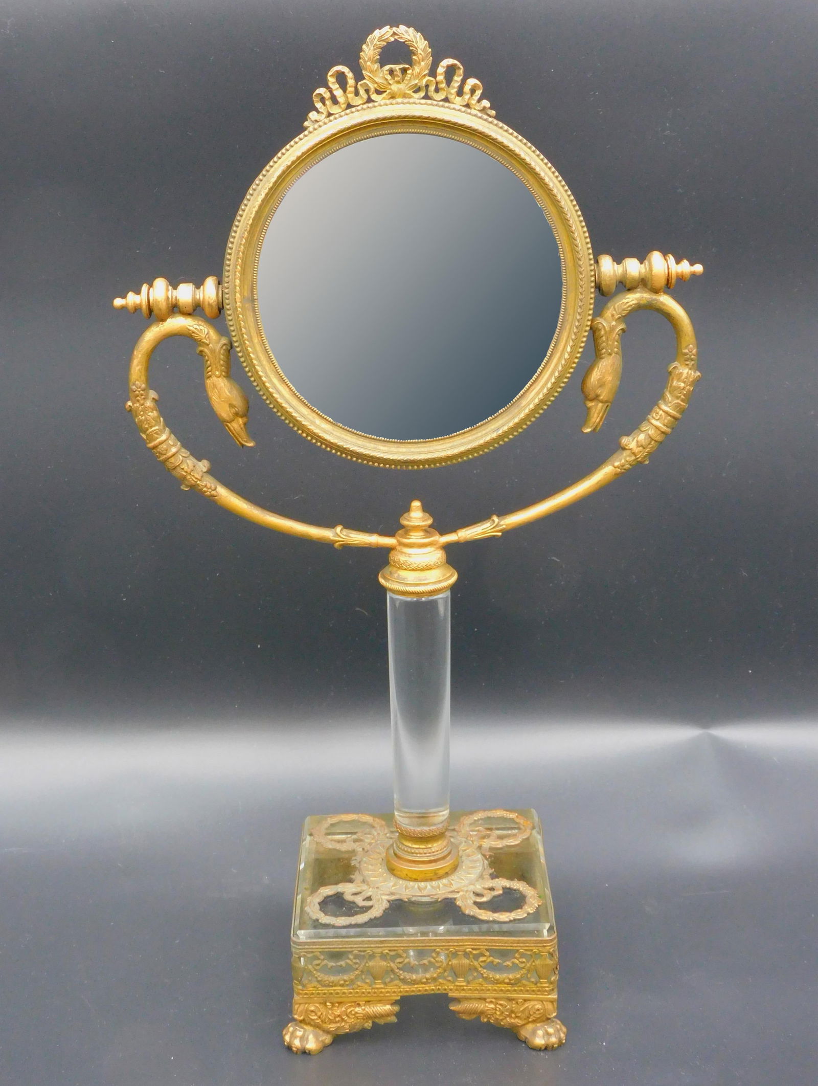 Double-sided French Neoclassical vanity mirror.: 19th century. Gilt bronze frame with crystal glass shaft and base. Decorated with swan heads, wreaths, garlands, urns, cornucopia, and animal pad feet. Beveled glass mirror. 15 1/2" high by 8 1/4" wid