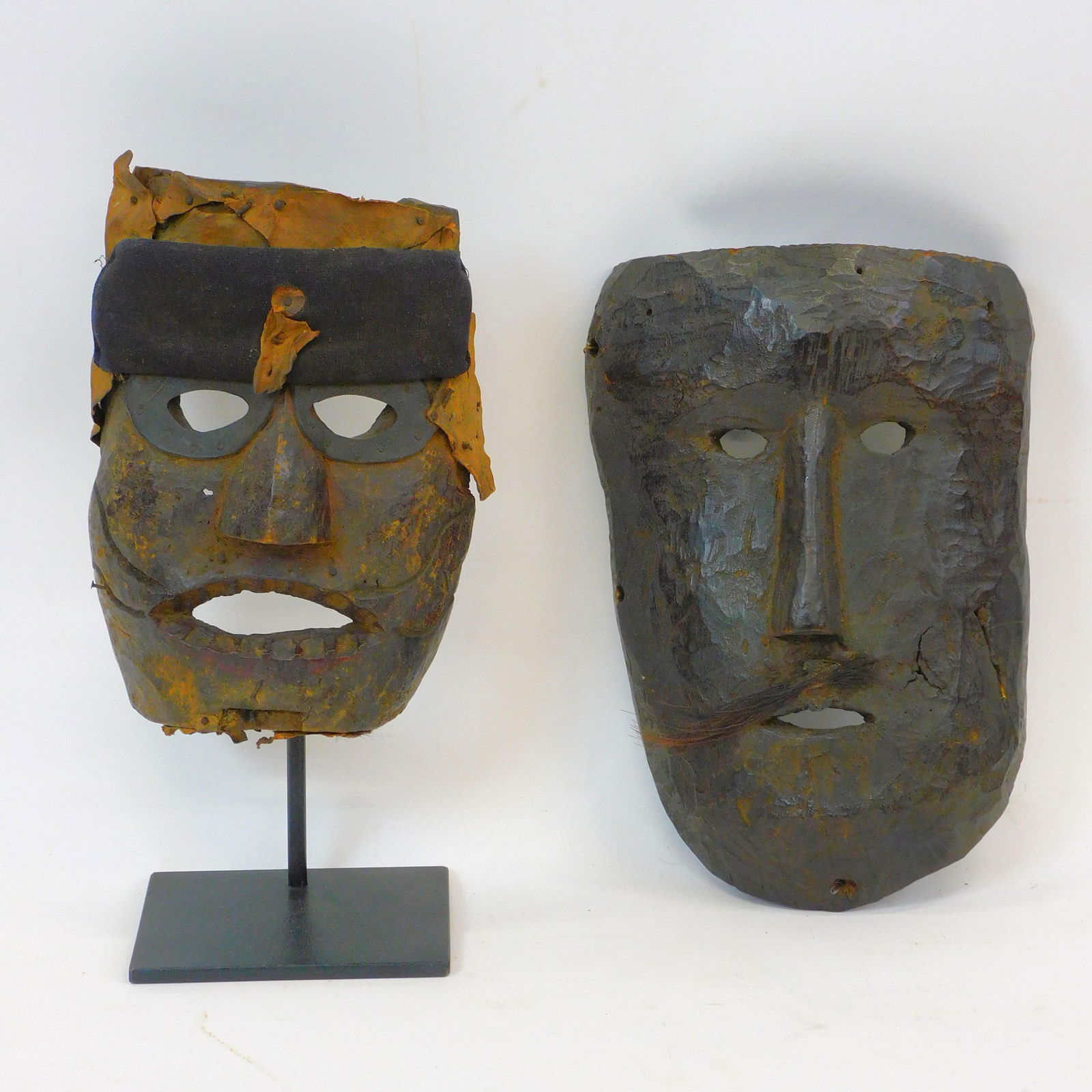 (2) Nepalese Shaman mask. Carved faces. Dark: natural finish. One has a mustache The other has applied leather decoration to one. One on museum mount stand. One is 11" by 7" wide. The other is 8" high by 6 1/4" wide. Wear consistent with age and
