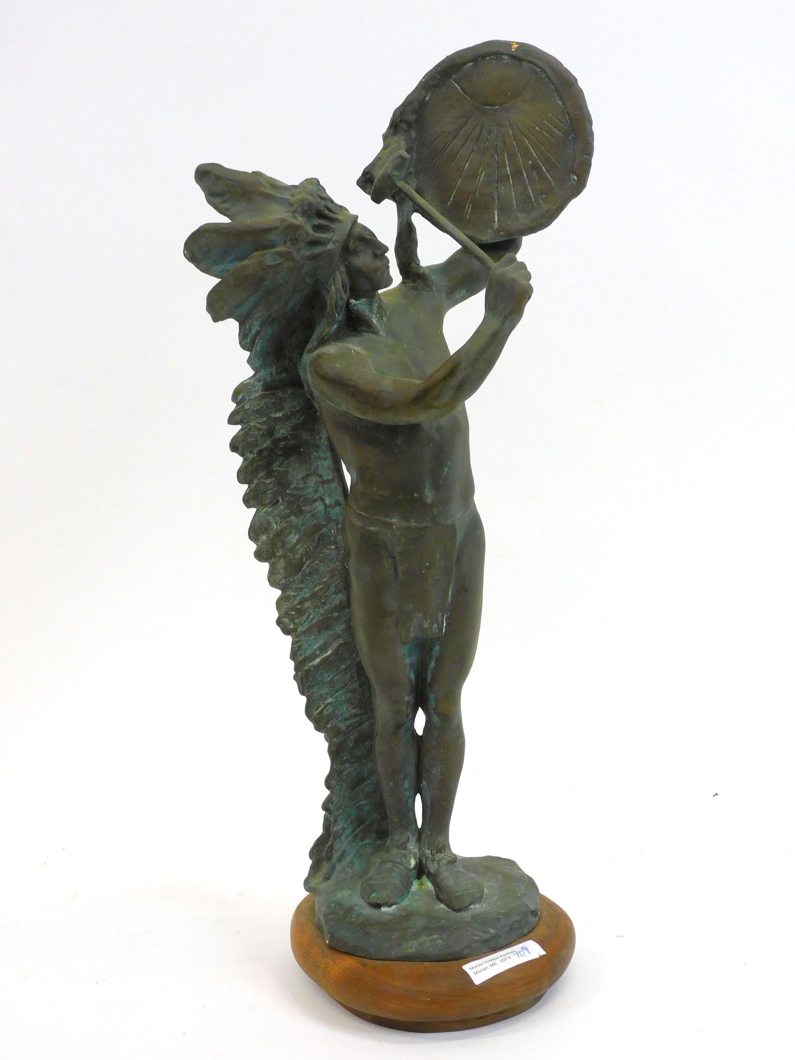 After Charles Henry Humphriss (1867-1934, New: York, England) bronze sculpture of a standing Native American Indian wearing a long feathered headdress. On a wooden base titled Sundial. The figure holds a large medallion to his side with a skull cr