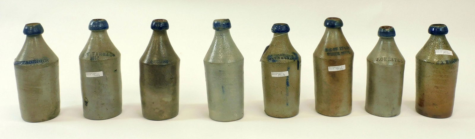 (8) Stoneware Bottles with Cobalt Enhancement.: 19th C. To include: J.A. Wallis, G. Vickary, M.H.S.W F.T. & Co White Beer, D.W. Tarr & Co 1852, Dennis & Gray 1854, J. Chester Lemon Beer, Tripp & Brig. CS, G.S. Allen. Roughly 9 1/2" to 10 1/2" high.