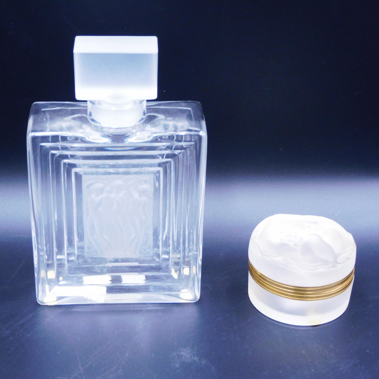(2) Lalique Crystal Pieces: To include: A) Covered dresser jar titled "Daphne" with image of young girl on lid. Signed in script "Lalique France". Brass trim. 2" high x 3" diameter. Good condition. B) Perfume bottle depicting th