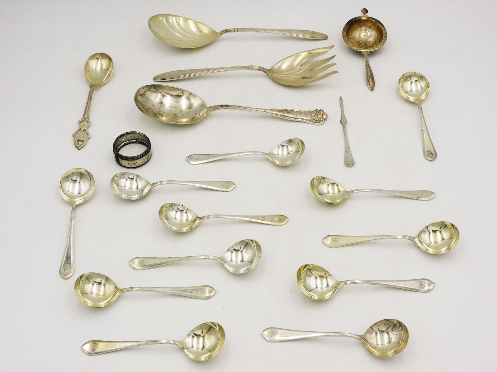 (18) Pieces Sterling Silver Flatware + Napkin Ring: To include: (A) Twelve bouillon spoons. Gorham. 5 1/4" long. (B) Two piece salad set. Gorham. 9" long. (C) Serving Spoon. Bigelow & Kennard. 9" long. (D) Sugar spoon. 6 1/4" long. (E) Tea strainer. 5