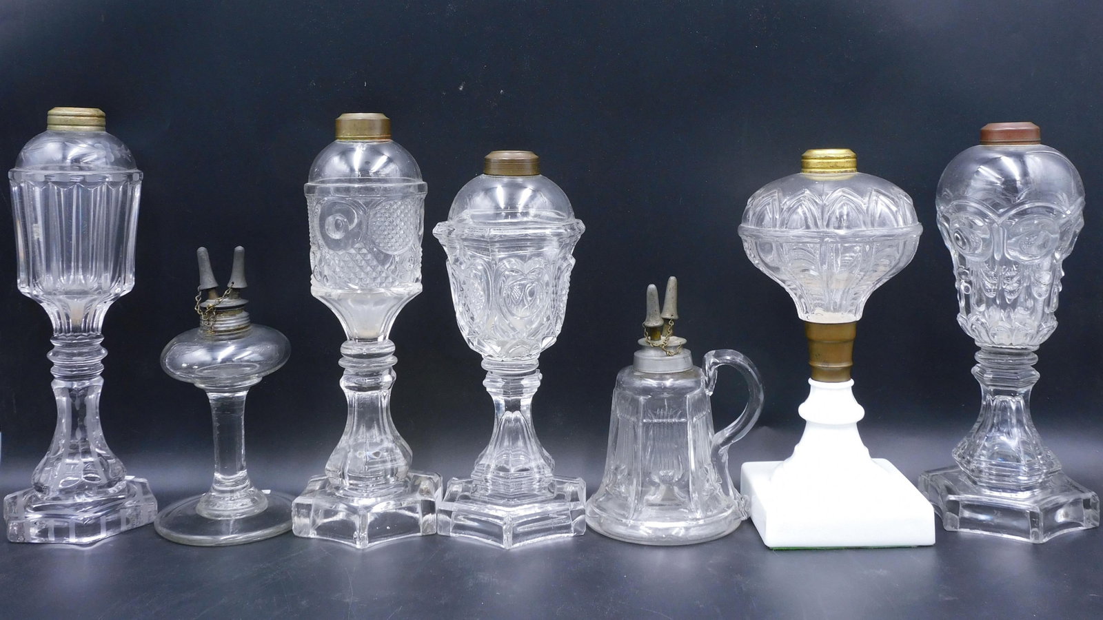 (7) Fluid Pattern Glass Lamps: 19th C. To include: (A) Bullseye and plume. 10 1/2" high. (B) Horn of plenty. 10 1/2" high. (C) Sweetheart. 9 1/4" high. (D) Pattern lamp with engraved design. 10 3/4' high. (E) Harp with camphene bur