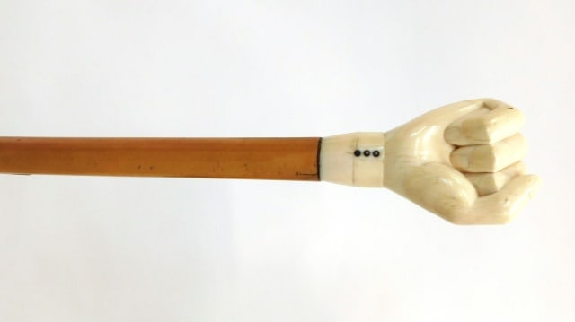 Scrimshaw Cane: 19th C. Tapered malaca wood shaft. Oversized pommel handle in the form of a clenched fist with a shirt cuff having three baleen buttons pinned with silver metal. 34" long. Old note on bottom tip: "Mrs