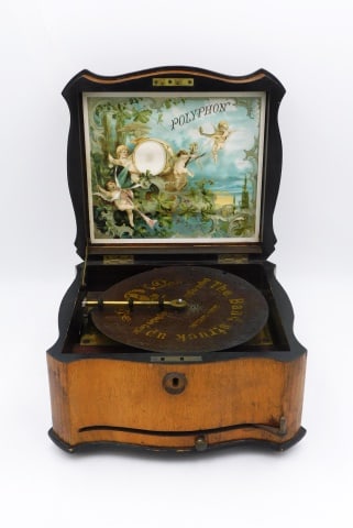 Polyphon Disc Music Box: Late 19th C. Mahogany case with inlaid top. Ratchet wind. Single comb. Original colored lithograph on lid depicting cherubs in a band. Comes with one disc. 6" high x 11" wide x 10" deep.