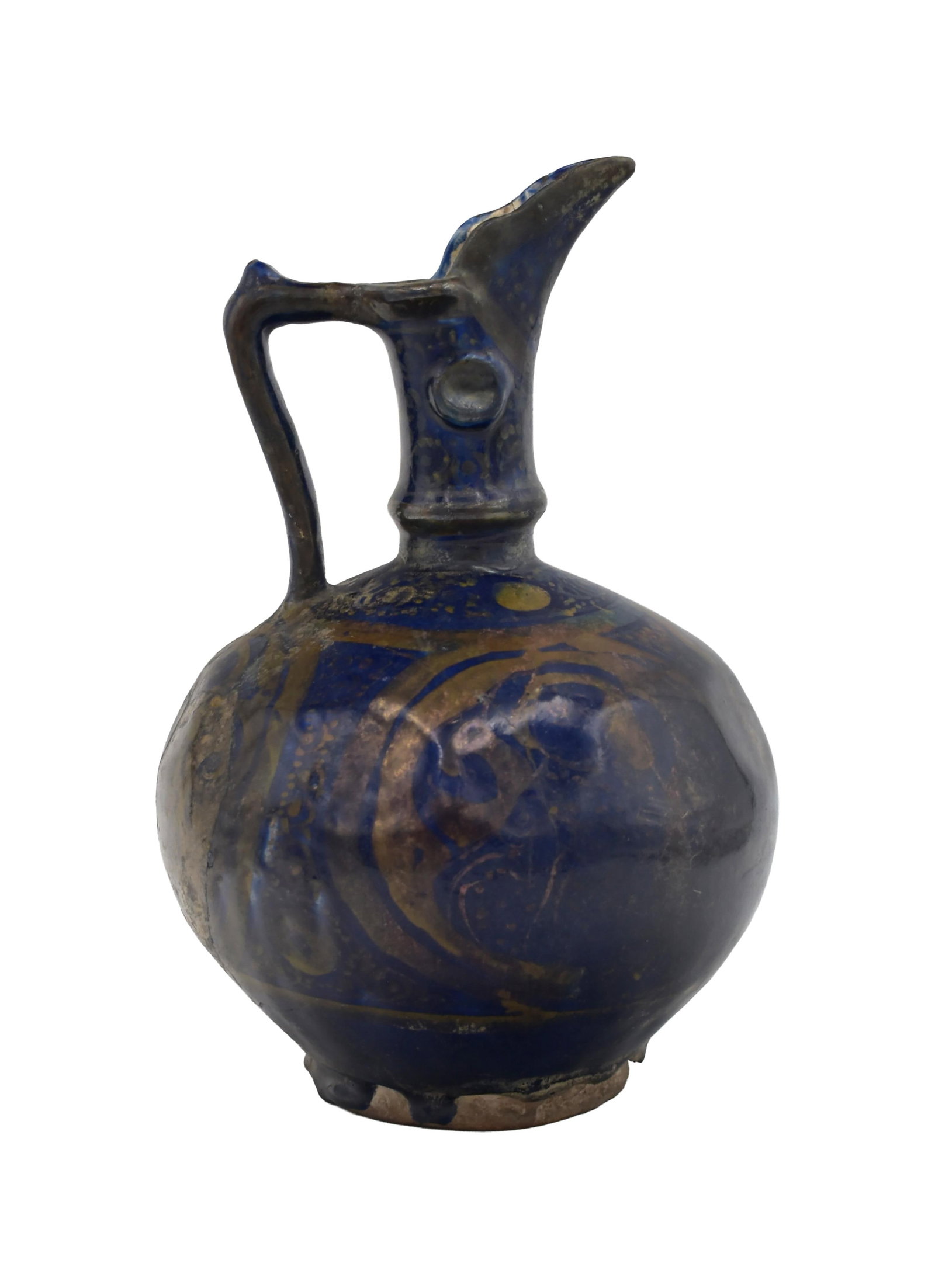 Persian Lustre Ewer: Mid-Late 12th C. Kashan or Rhages. Ceramic ewer with an indigo glaze. Beak-style spout. Shouldered handle. Globular body. Nodules on the neck are in the style of bronzes of the same era. Painted