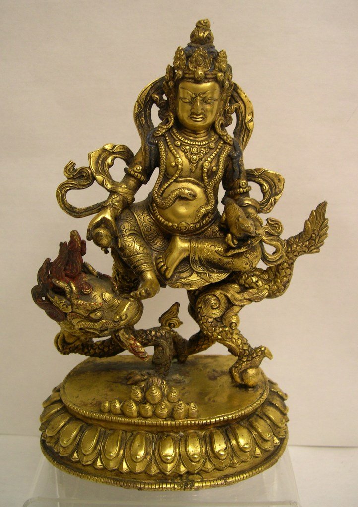 Gilt Bronze Deity of Dharmapal and Dragon.: Head with chignon and tiara, flowing ribbon around body, beaded jewelry with snake on neck, right hand with rat, left hand a magic ball, crossed left leg, right leg resting on dragon. Red pigments to 