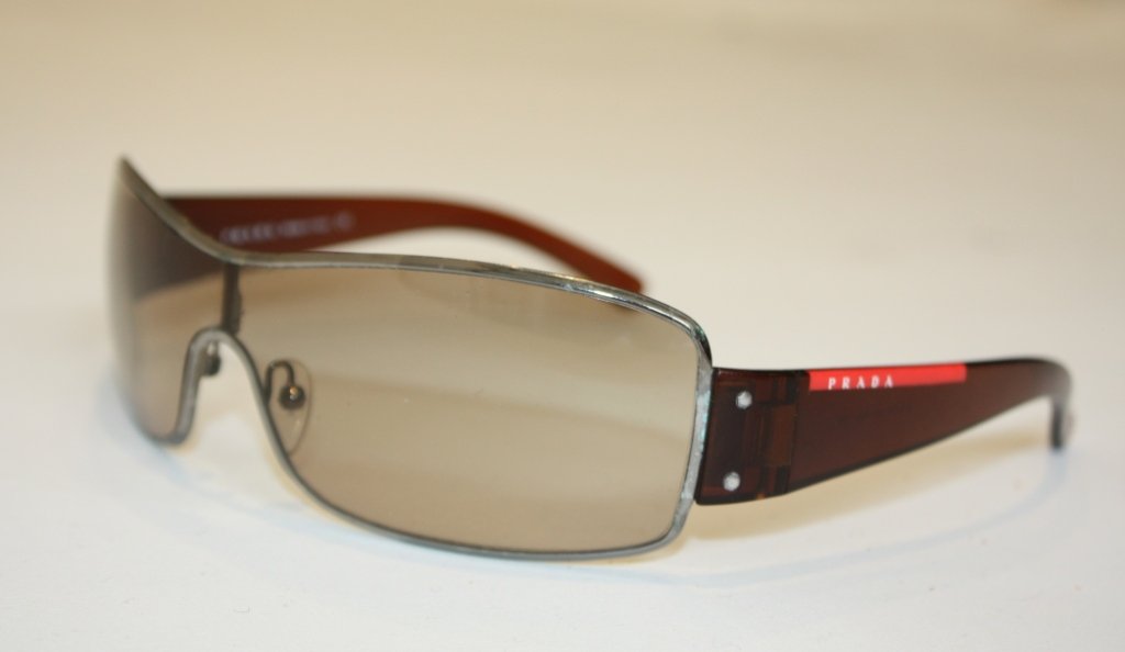 PRADA: PRADA Paire de lunettes, branches en acétate marron siglées. Dans son étui en pvc rouge.