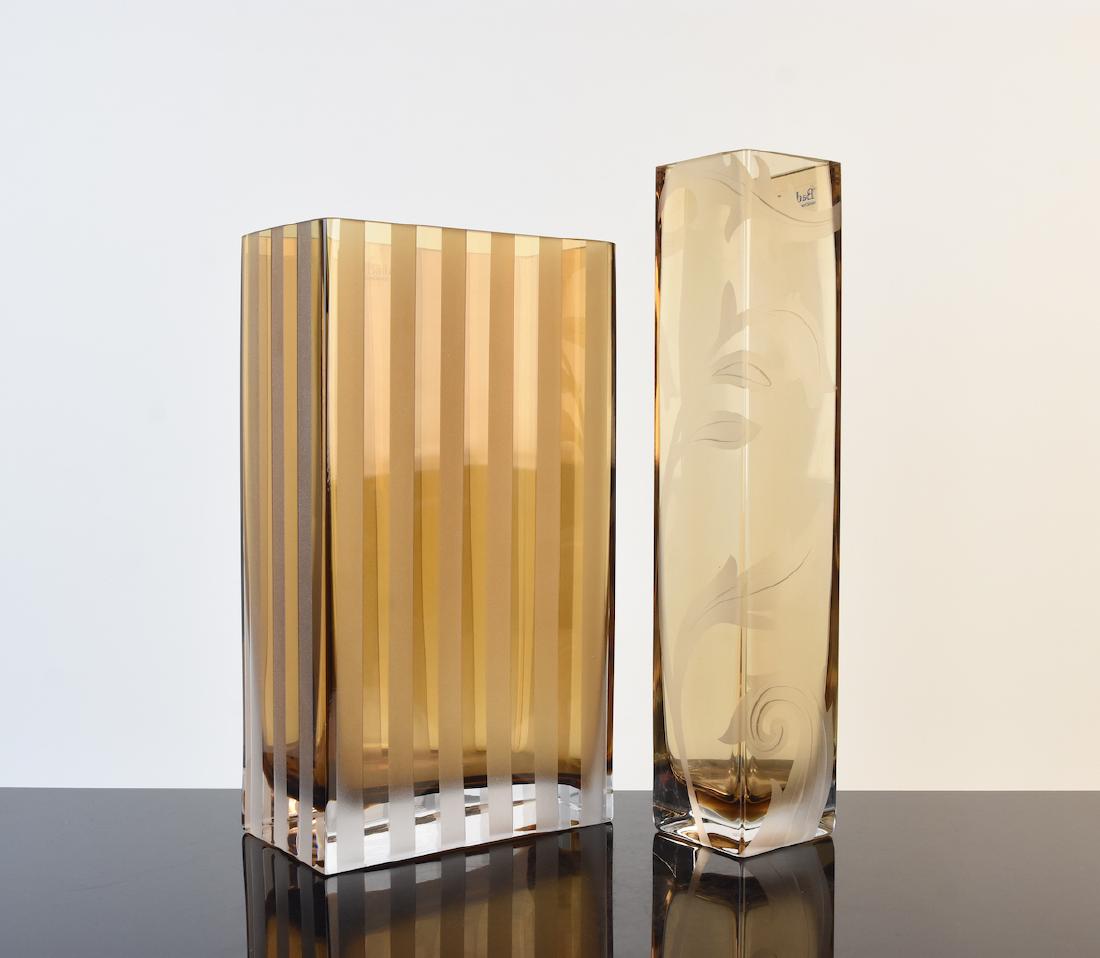Complementary Badash Crystal Vases: Badash hand made crystal vases in an amber hue. One vase is square shaped with frosted leaves adorning two sides. The second vase is rectangular with frosted pinstripes on each side. Both vases are in
