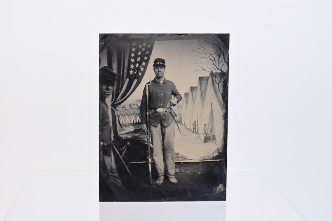 Civil War Union Soldier Tintype 1/4 Plate: A 1/4 plate tintype (ferrotype) photograph of a Union soldier from the Civil War. The gentleman is standing in front of a clock backdrop depicting barracks and a second man can be seen at the left. He