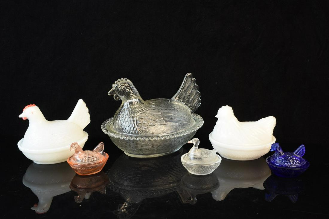 Glass Hen on Nest Dishes: An assortment of six glass nesting hen and duck dishes in various sizes. Includes clear pressed glass; milk glass; pink tinted glass; and cobalt blue glass. Light surface markings. No discernible make