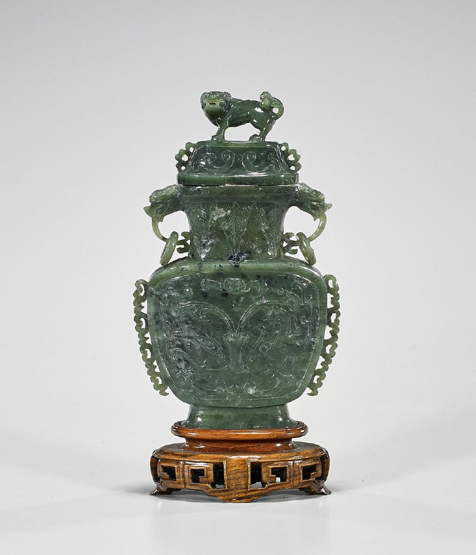 Chinese Carved Spinach Jade Covered Urn: Chinese carved spinach jade urn; of flattened form, with animal handles pending faux rings; the body showing taotie mask designs flanked by openwork protruding flanges; surmounted by a cover with lion