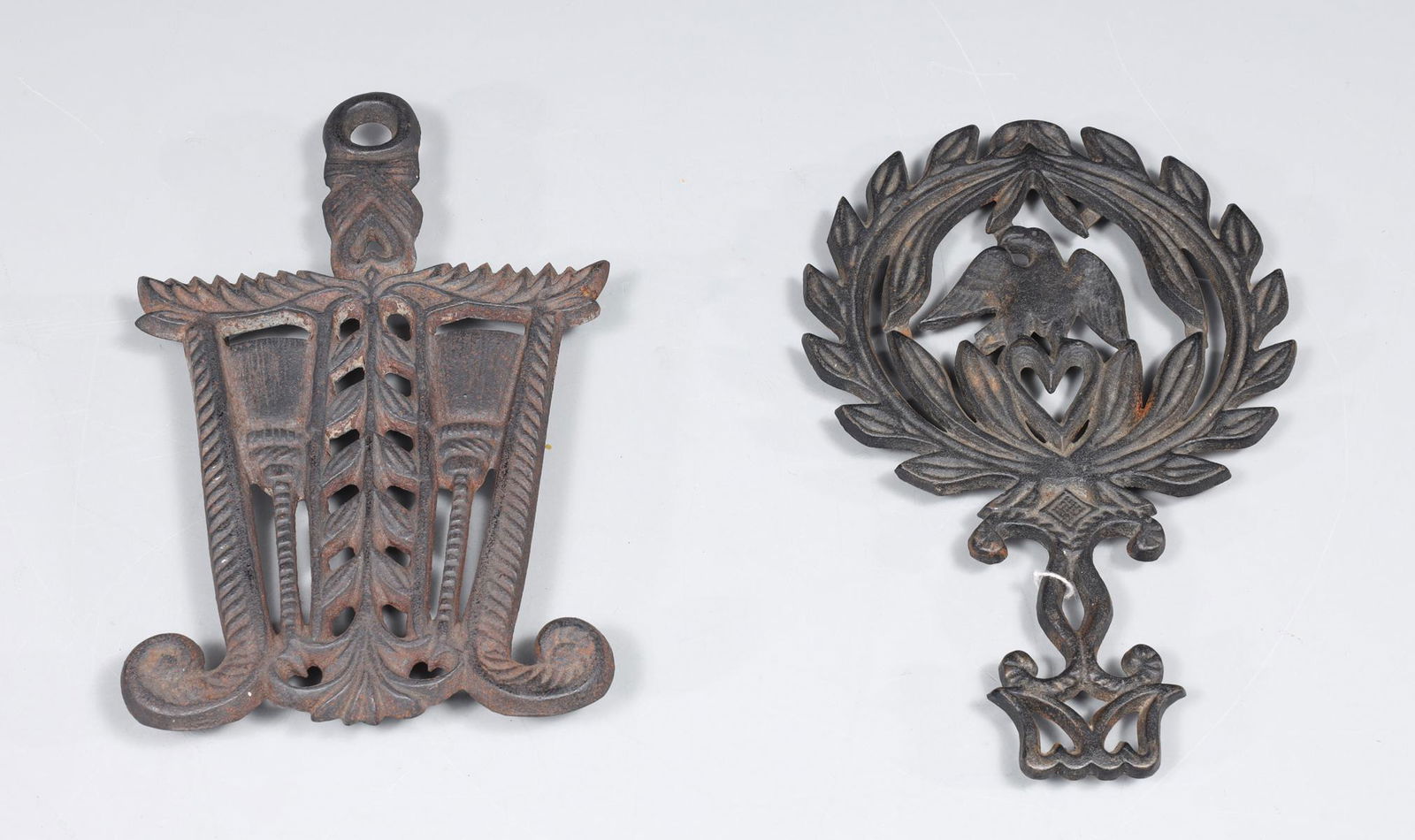 Group of Two Antique Cast Iron Trivets: Group of two antique cast iron trivets, John Wright Co. trivet with tassel detail: patina, moderate age wear, surface soiling; Wilton eagle and laurel motif: patina, age wear; largest L 5" W 8 1/2" H