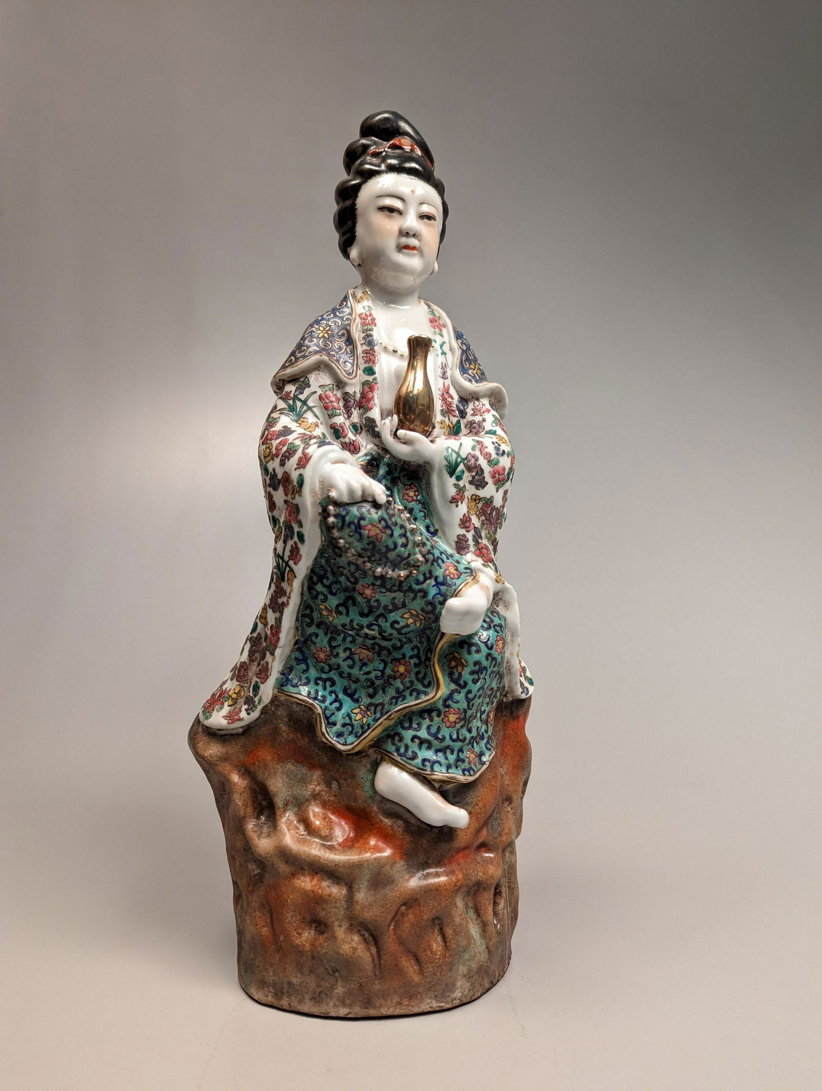 Chinese Enameled Porcelain Guanyin: Chinese enameled porcelain figure; of Guanyin seated atop a tall rockery base; holding her bottle of tears in one hand, and a strand of prayer beads in the other; dressed in ornate floral robes; minor