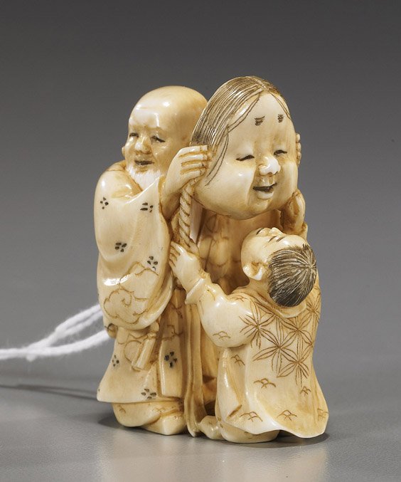 Carved Ivory Netsuke: Figures with Mask: Carved Ivory Netsuke: Figures with Mask Well carved and lightly stained, ivory netsuke of 2 figures: a man and boy holding a large mask of Okame, signed; Provenance: purchased from Michael Spindel in 