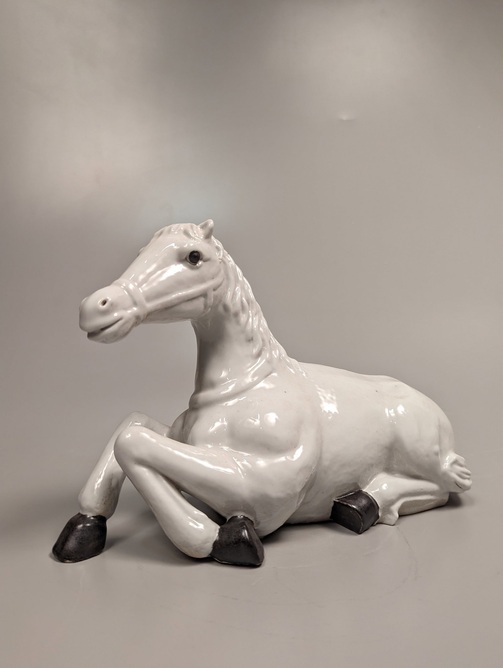 Chinese Republic-Style Glazed Porcelain Horse: Chinese Republic-style white glazed porcelain of a horse in repose, with modeled trappings, and black glazed hooves; usual minor wear; L: 12 1/2" (approx.)