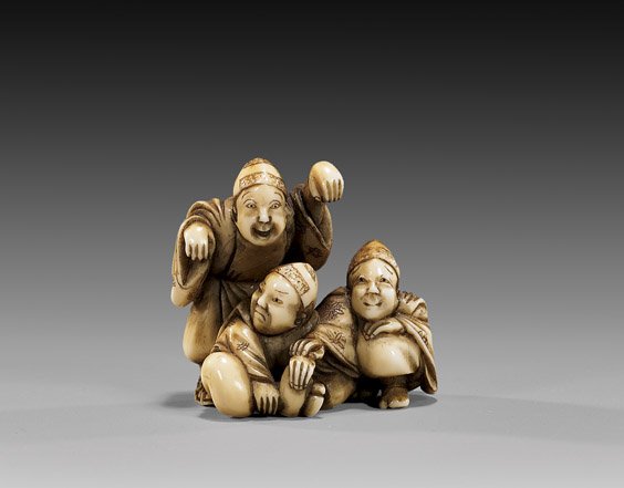 ANTIQUE IVORY NETSUKE: Three Figures: 象牙根付 ANTIQUE IVORY NETSUKE: Three Figures Antique carved ivory netsuke group of three figures: two seated and one standing in animated pose; signed Tomokazu, 19th Century; 