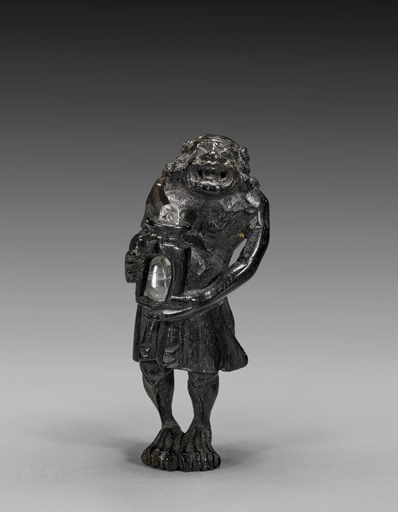 ANTIQUE WOOD NETSUKE: Oni: 木製根付 ANTIQUE WOOD NETSUKE: Oni Antique carved wood netsuke; of an oni standing and holding a small scholar's table/stand; set with later rock crystal quartz; early 19th Cen
