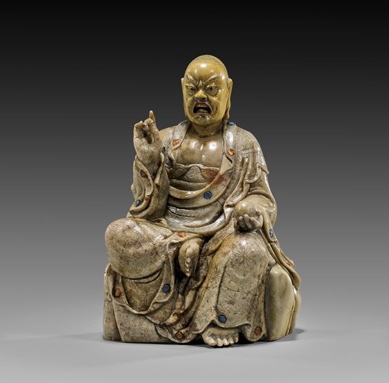 CHINESE CARVED SOAPSTONE LOHAN: CHINESE CARVED SOAPSTONE LOHAN Dramatically carved, Chinese soapstone figure; of a seated Lohan in the position of "Royal Ease" upon rockery, wearing an unusual facial expression and set with small ca