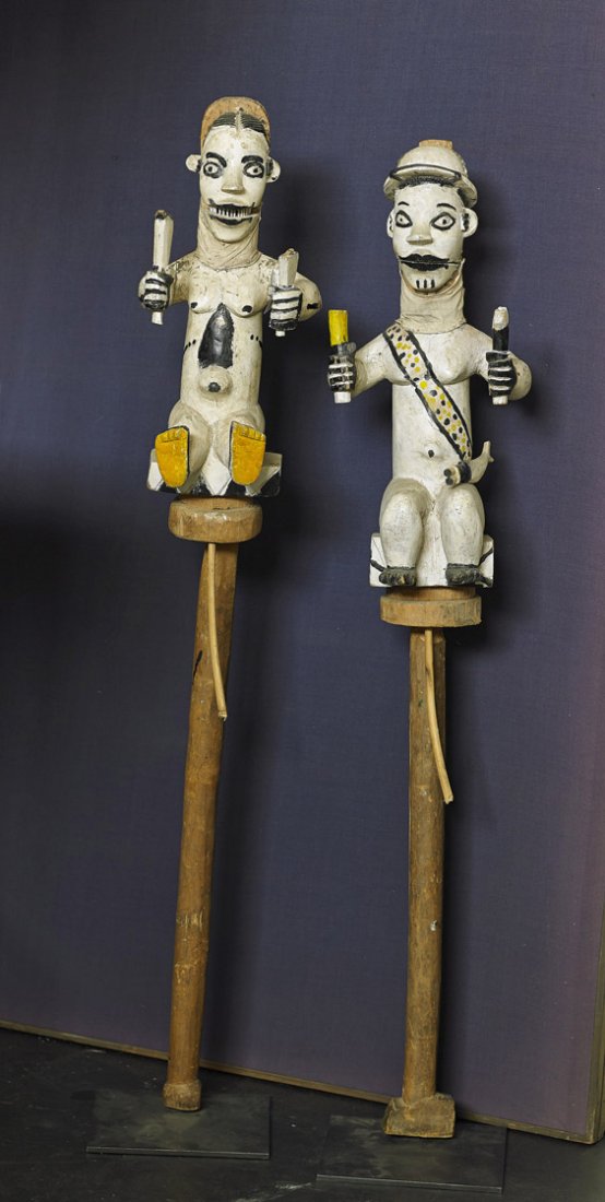 Pair African Carved Wood Figural Puppets: Pair African Carved Wood Figural Puppets Pair tall African carved and painted wood figural puppets; each seated and holding objects in each hand: both wearing a sash, one with a cap, both atop a long 