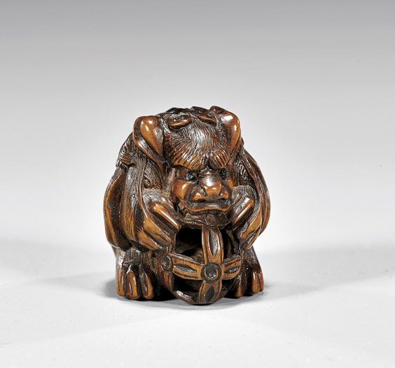 CARVED WOOD NETSUKE: Shi-Shi: 木彫根付 CARVED WOOD NETSUKE: Shi-Shi Carved wood netsuke; of Shi-shi hunched over an openwork ball, containing a rattling ball inside