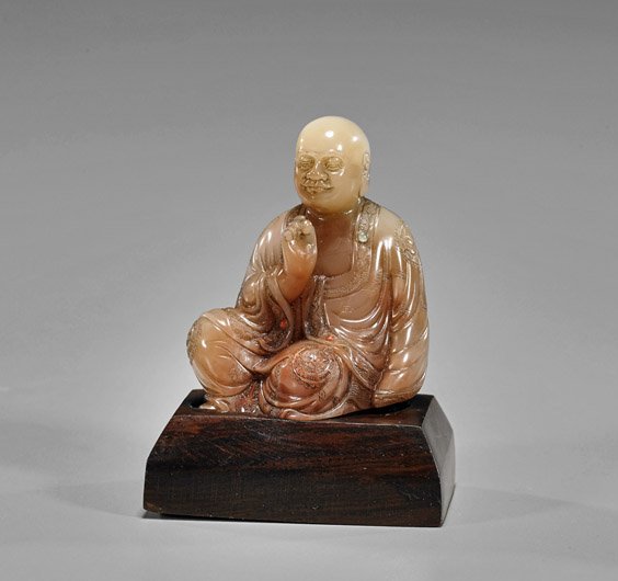 Small Chinese Carved Soapstone Lohan: Small Chinese Carved Soapstone Lohan Small Chinese carved soapstone Lohan; the sitting deity in "Royal Ease" with decorative robes and inlaid gems, of pale red suffusions; H: 2 1/2"; wood stand
