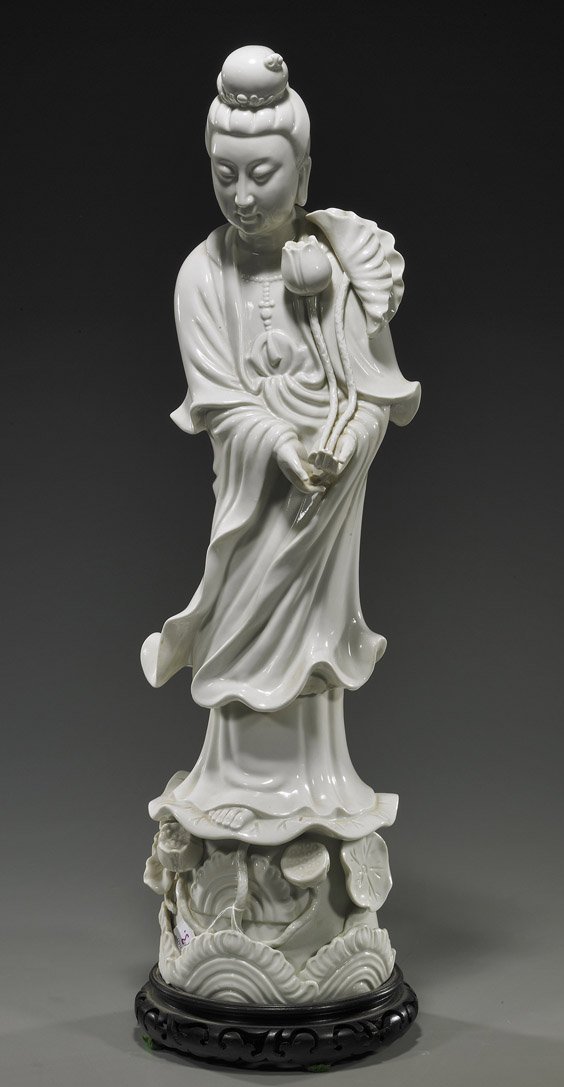 Tall Blanc de Chine Porcelain Guanyin: Tall Blanc de Chine Porcelain GuanyinTall Chinese, blanc de chine porcelain model of Guanyin; standing in long robes atop a lotus pedestal and carrying a lotus flower; H: 23 1/2", wood stand