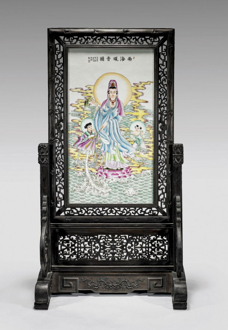 FAMILLE ROSE PORCELAIN TABLESCREEN: Chinese Famille Rose enameled porcelain tablescreen; the panel depicting a Guanyin with a child to either side, standing upon a lotus base upon a ground of waves; in openwork carved wood stand and fra