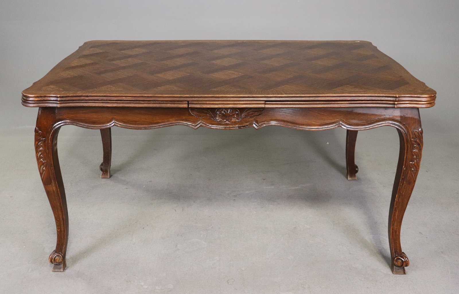 French Louis XV Style Oak Parquetry Dining Table: French Louis XV style oak dining table featuring a decorative parquetry top in a geometric basket-weave pattern. Shaped apron with carved floral accents raised on cabriole legs with carved details and